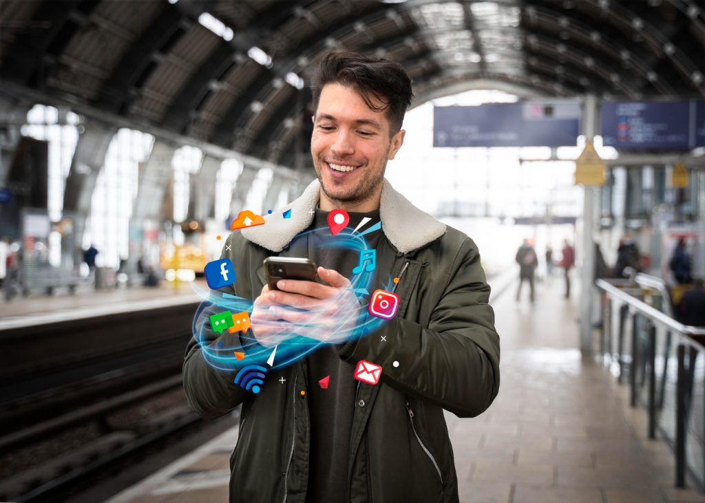 Young man at a Brisbane train station smiling at his phone with digital marketing and social media icons floating around, symbolizing online marketing and social media growth for Brisbane businesses.